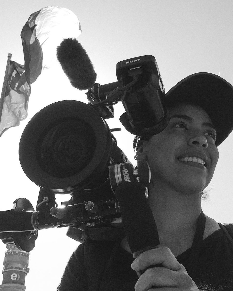 Jessica Villamil en rodaje, sonriendo junto a una cámara con óptica ARRI; al fondo, una bandera de México y el cielo a contraluz.