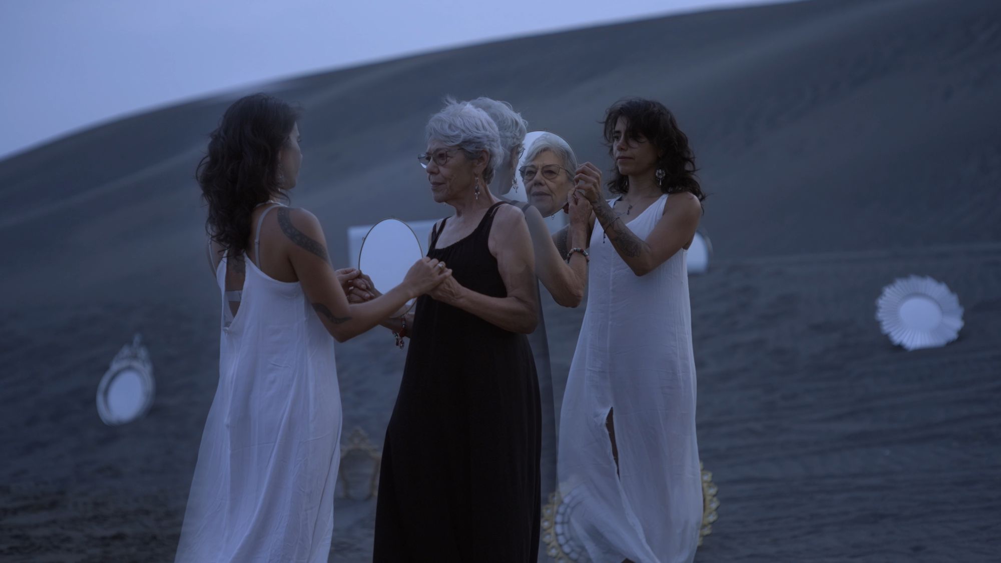 Cuatro mujeres vestidas de blanco y negro sobre la arena, rodeadas de espejos.