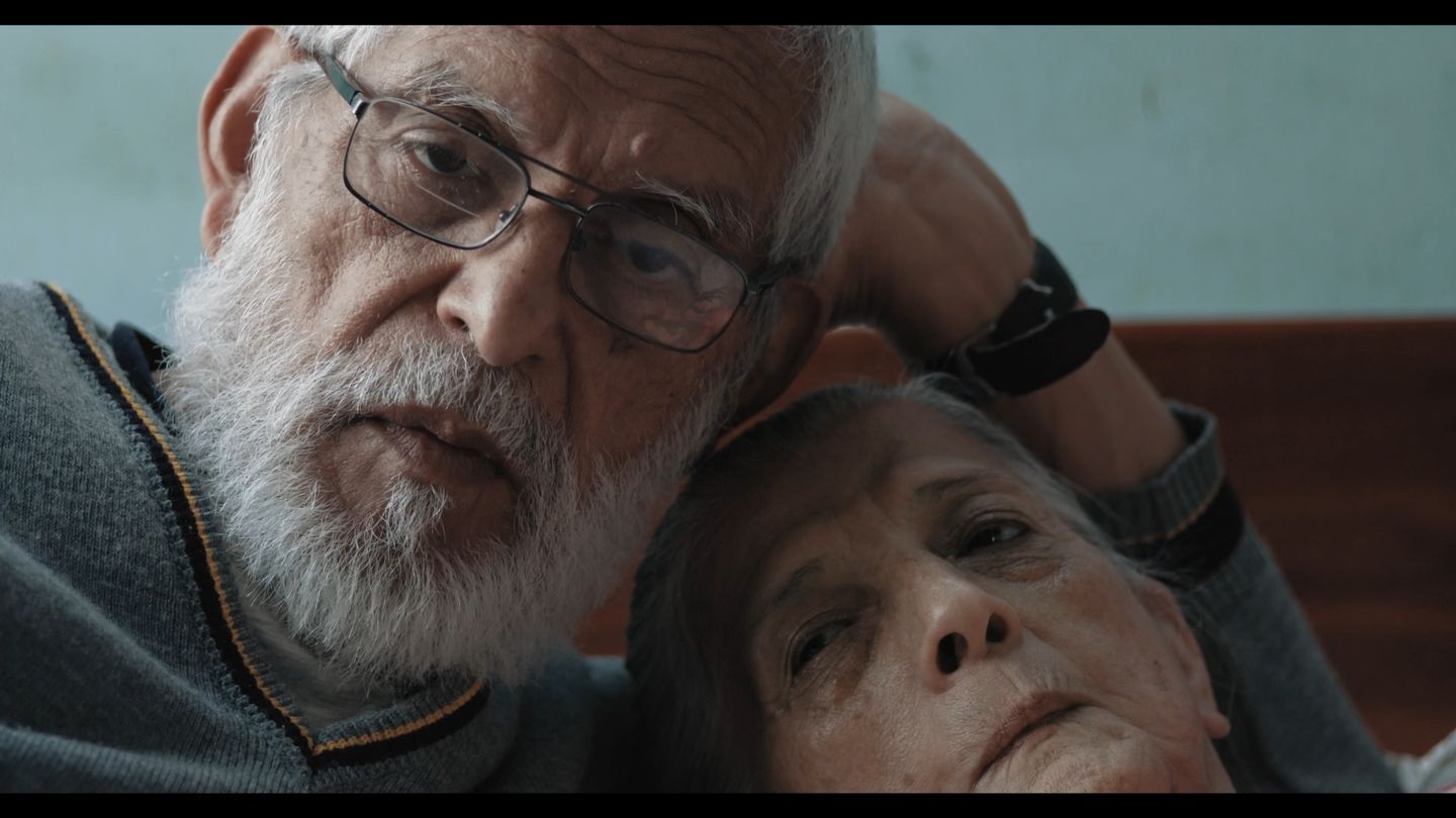 Un hombre de barba blanca y lentes sostiene con ternura la cabeza de su compañera, muy cerca de la suya.