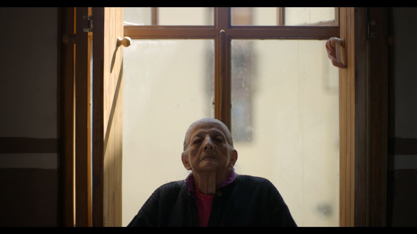 Una mujer mayor se asoma al marco de una ventana, recortada por la luz.