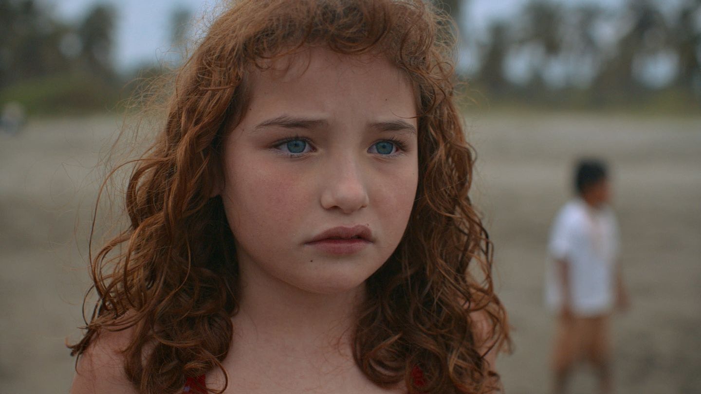 Retrato cercano de una niña en la playa; ojos claros y cielo velado al fondo.