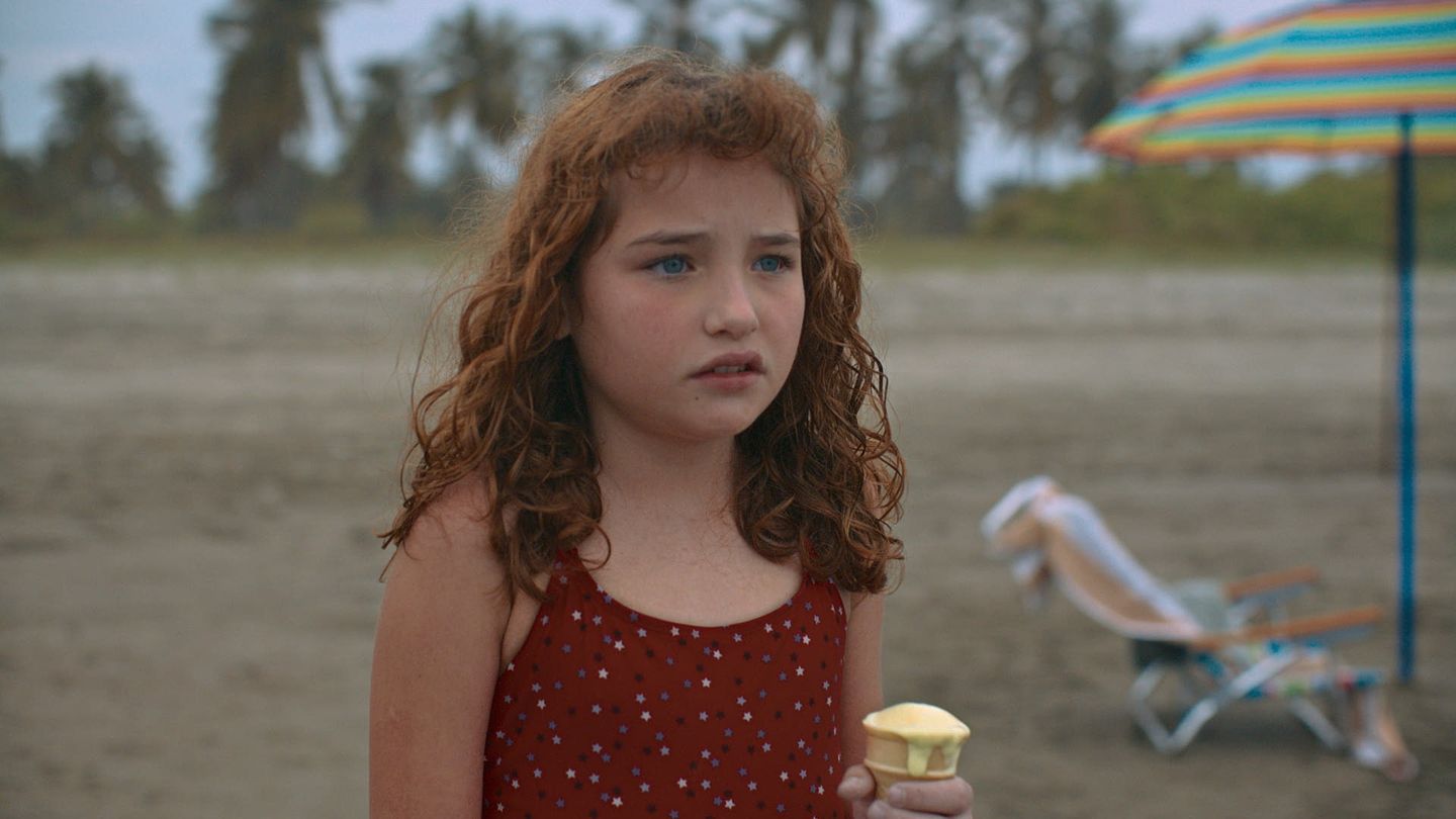 Una niña con traje de baño rojo camina por la playa junto a una sombrilla.